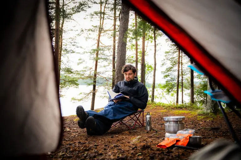 Uitzicht vanuit de tent op een man die comfortabel leest op een afgelegen wildkampeerplek aan een meer in het Zweedse Värmland, omgeven door dennenbomen.