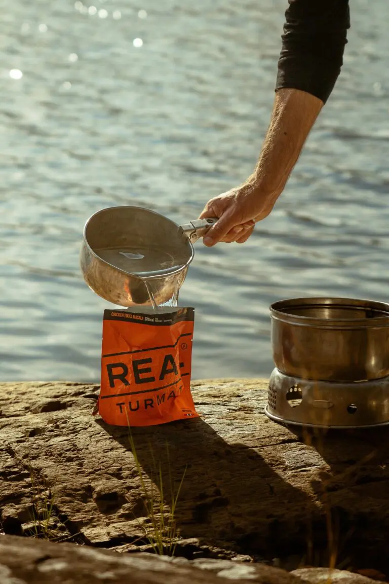 person pouring water into a dehydrated meal pouch by a lake
